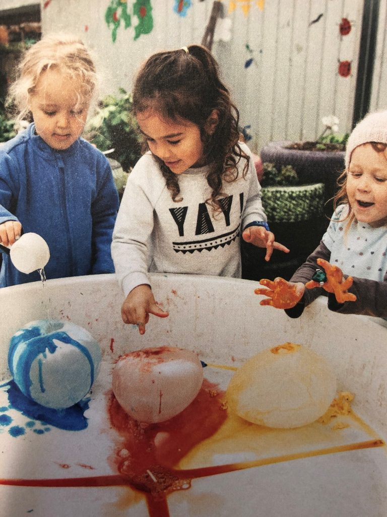 Three young children playing with large balls of ice, using paint and water to experiment with them.